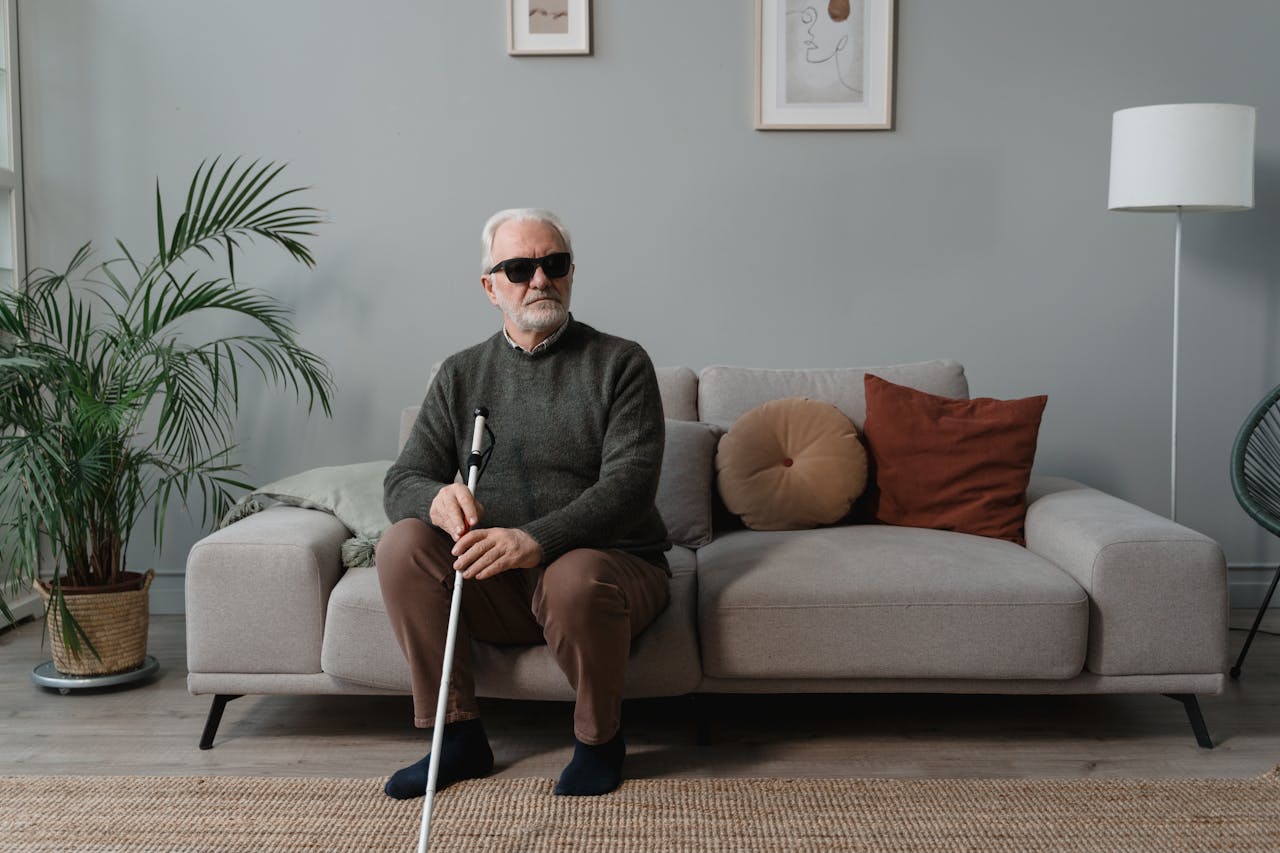 Elderly man with sunglasses and white cane sitting on a sofa, symbolizing independence and support.