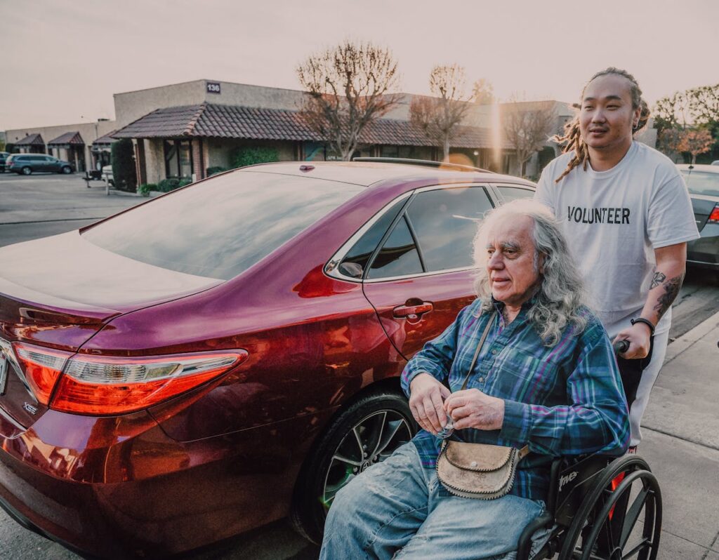 A volunteer helps an elderly man in a wheelchair near parked cars on a sunny day.